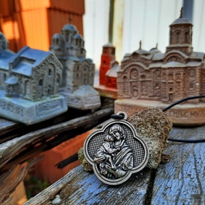 May include: A silver pendant with a depiction of the Virgin Mary holding a baby Jesus. The pendant is hanging from a black cord and is in front of several small, detailed model churches.