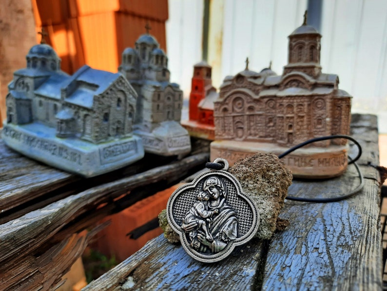 May include: A silver pendant with a depiction of the Virgin Mary holding a baby Jesus. The pendant is hanging from a black cord and is in front of several small, detailed model churches.