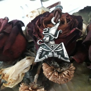 Puede incluir: Collar con colgante de plata que representa a un dios egipcio con un tocado y sosteniendo un bastón. El colgante está en un cordón de cuero marrón y está rodeado de rosas secas.