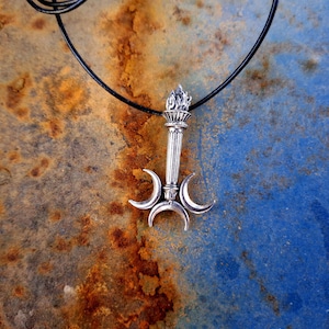 Op de afbeelding: Zilverkleurige hanger ketting met een fakkel ontwerp, vlammen aan de bovenkant en drie halve maan vormen eronder. De hanger hangt aan een zwart koord tegen een getextureerde, roestige blauw-bruine achtergrond.