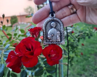 May include: Silver pendant necklace with a religious icon of a person holding a book and a staff. The pendant is in a silver frame with a decorative border. The pendant is hanging from a black leather cord.