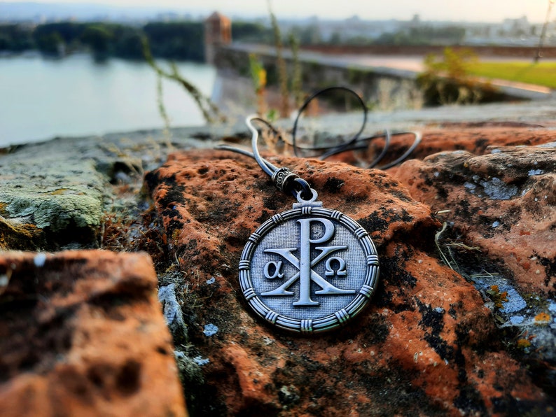 May include: A silver pendant necklace with a round charm featuring the Chi Rho symbol, alpha and omega, and the letters 'P' and 'X'. The necklace is on a brown and green rocky surface.
