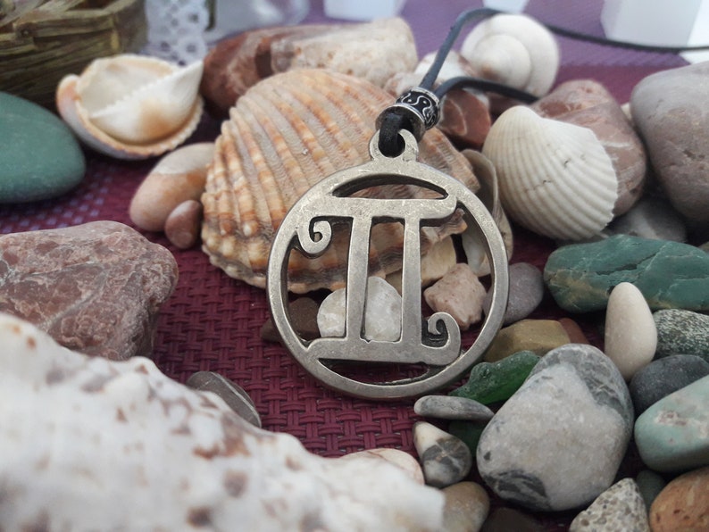 May include: A silver necklace with a circular pendant featuring the Gemini zodiac symbol. The pendant is surrounded by various seashells and stones.