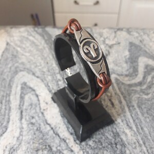 May include: A brown leather bracelet with a silver charm featuring the astrological symbol for Aries. The bracelet is displayed on a black stand.