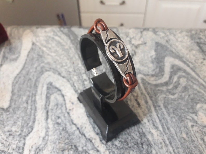 May include: A brown leather bracelet with a silver charm featuring the astrological symbol for Aries. The bracelet is displayed on a black stand.