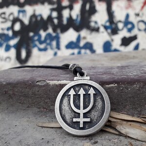 May include: A silver pendant necklace with a circular design featuring a trident and a cross. The pendant is on a black cord and is resting on a gray surface.