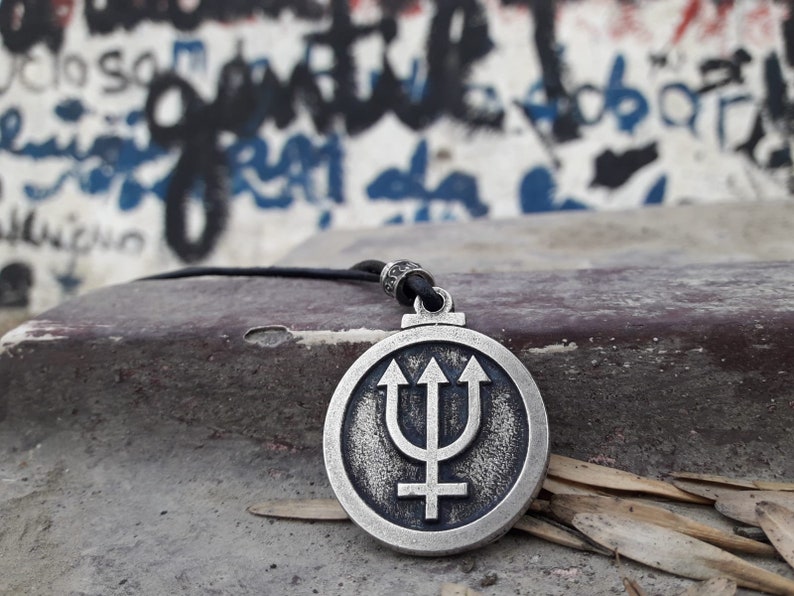 May include: A silver pendant necklace with a circular design featuring a trident and a cross. The pendant is on a black cord and is resting on a gray surface.
