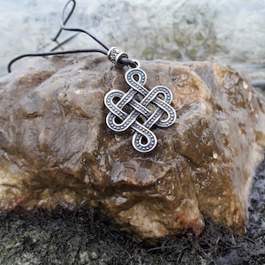 May include: A silver Celtic knot pendant necklace with a black leather cord. The pendant is a complex design of interwoven knots, and it is resting on a grey rock.