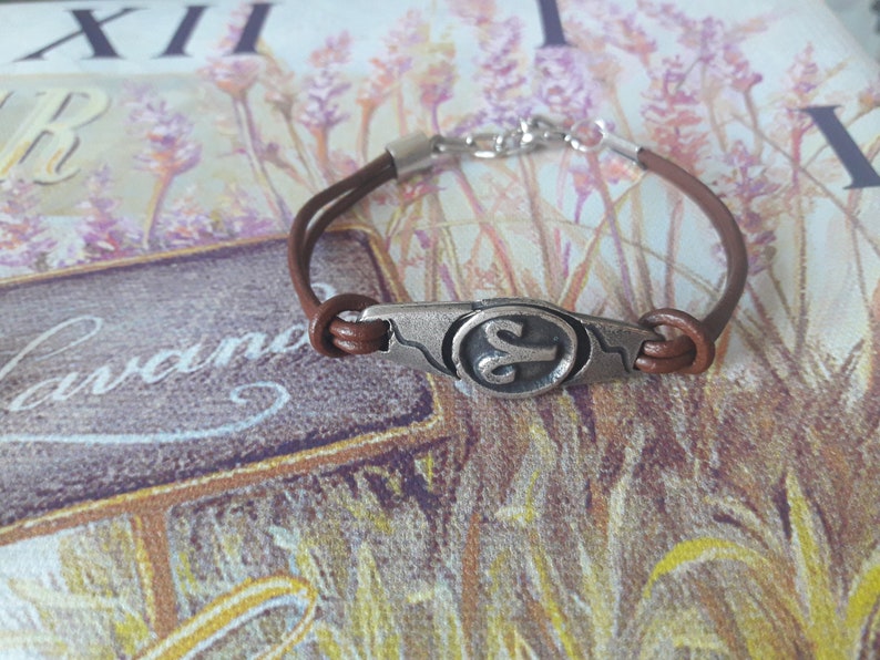May include: Brown leather bracelet with a silver charm featuring the astrological symbol for Aries.