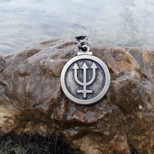May include: A silver pendant necklace with a circular design featuring a trident and a cross. The pendant is on a black cord and is resting on a brown rock.
