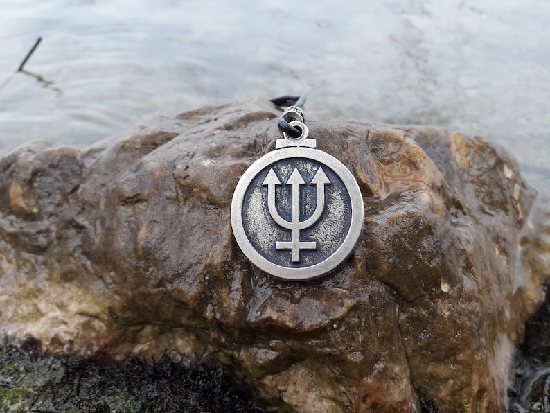 May include: A silver pendant necklace with a circular design featuring a trident and a cross. The pendant is on a black cord and is resting on a brown rock.