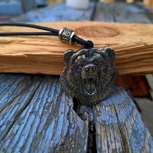 May include: A silver bear head pendant with an open mouth and sharp teeth, hanging from a black leather cord with a silver bead.