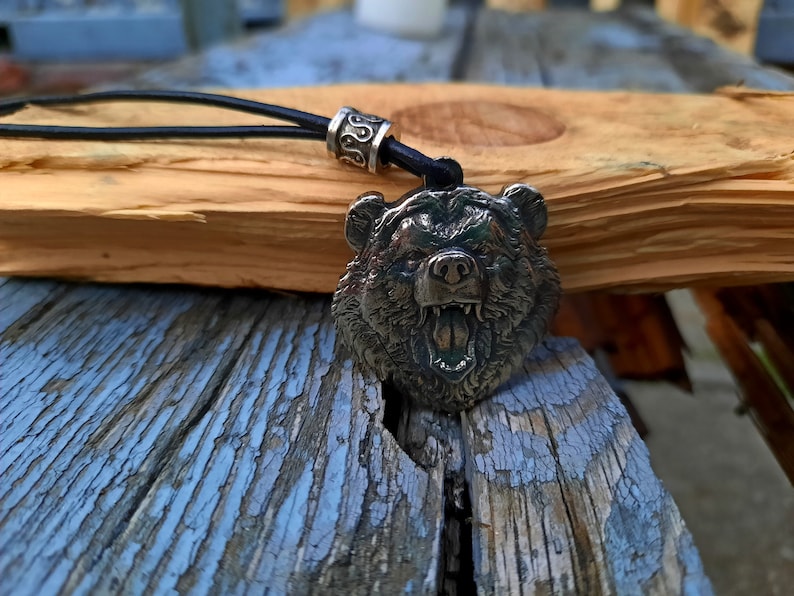 May include: A silver bear head pendant with an open mouth and sharp teeth, hanging from a black leather cord with a silver bead.