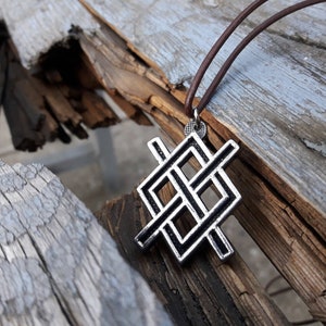 May include: Silver-tone pendant necklace with a geometric design, on a brown leather cord. The pendant has a black inlay, set against a weathered wooden background.