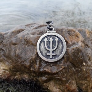 May include: A silver pendant with a trident and a symbol resembling a cross, set against a circular background. The pendant is resting on a brown rock.