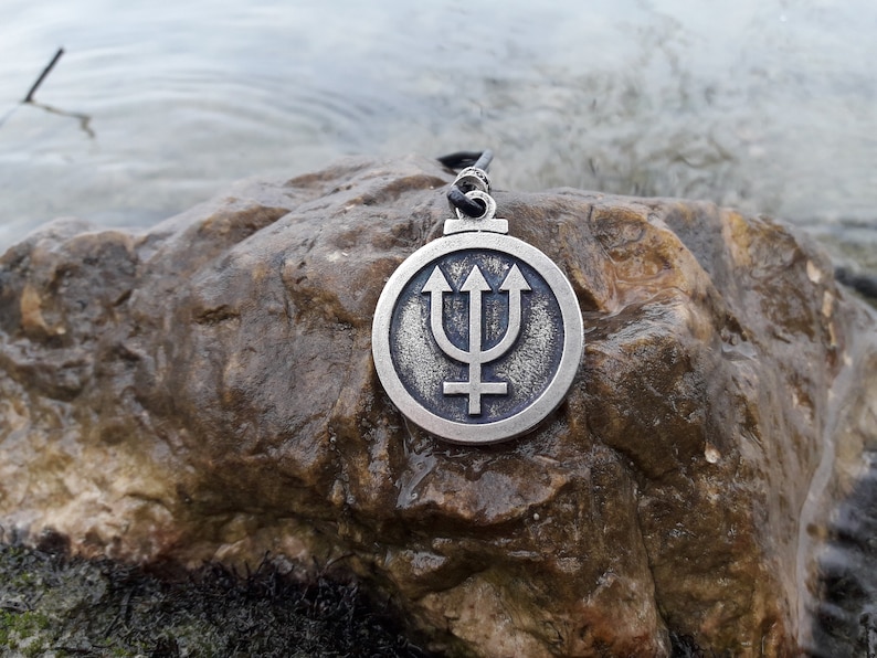 May include: A silver pendant with a trident and a symbol resembling a cross, set against a circular background. The pendant is resting on a brown rock.