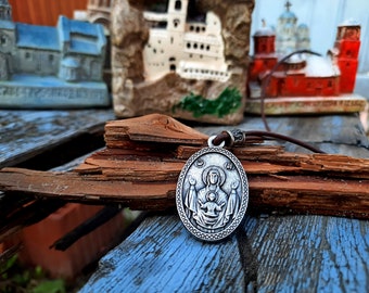 May include: Oval silver pendant featuring a religious icon of a woman and child, suspended from a brown leather cord. The pendant has a detailed relief design and is set against a rustic wooden background.