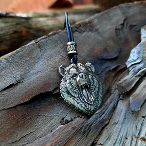May include: A silver bear head pendant necklace with a black cord. The bear's mouth is open and its teeth are visible. The pendant is on a wooden surface.