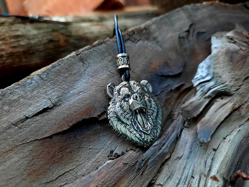 May include: A silver bear head pendant necklace with a black cord. The bear's mouth is open and its teeth are visible. The pendant is on a wooden surface.