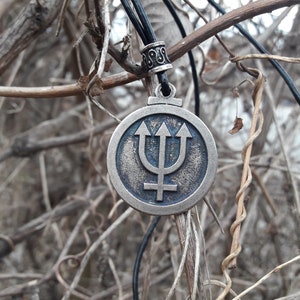 May include: A silver-toned pendant necklace with a round charm featuring a blue trident symbol and a stylized planet symbol. The necklace is hanging from a black cord.