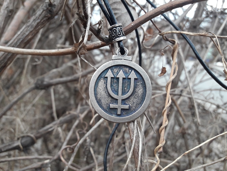May include: A silver-toned pendant necklace with a round charm featuring a blue trident symbol and a stylized planet symbol. The necklace is hanging from a black cord.