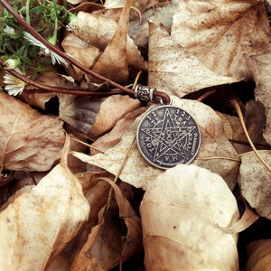 May include: A silver pendant with a five-pointed star and the words "TETRAGRAMMATON" and "123456789101112" around the edge, resting on a bed of brown leaves.