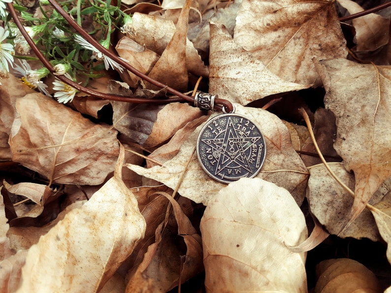 May include: A silver pendant with a five-pointed star and the words "TETRAGRAMMATON" and "123456789101112" around the edge, resting on a bed of brown leaves.