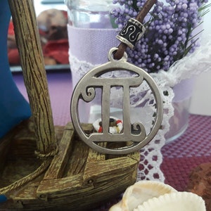 May include: Silver pendant necklace with a Gemini zodiac symbol charm. The charm is round and features a detailed design of the Gemini symbol. The necklace is on a brown leather cord with a silver bead.