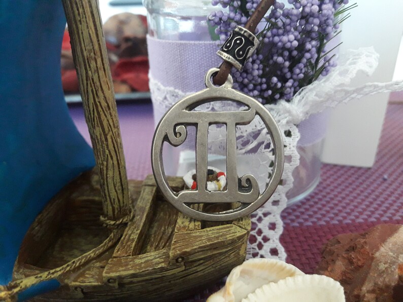 May include: Silver pendant necklace with a Gemini zodiac symbol charm. The charm is round and features a detailed design of the Gemini symbol. The necklace is on a brown leather cord with a silver bead.
