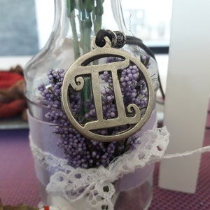 May include: A silver pendant necklace with a circular charm featuring the Gemini zodiac symbol. The charm is surrounded by purple flowers and white lace.