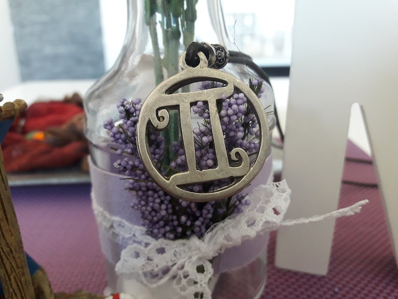 May include: A silver pendant necklace with a circular charm featuring the Gemini zodiac symbol. The charm is surrounded by purple flowers and white lace.