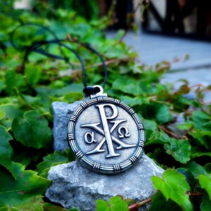 May include: A silver pendant necklace with a round, ornate design featuring the Chi Rho symbol, alpha and omega, and the letter P. The pendant is on a gray rock with green foliage in the background.