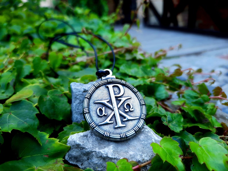 May include: A silver pendant necklace with a round, ornate design featuring the Chi Rho symbol, alpha and omega, and the letter P. The pendant is on a gray rock with green foliage in the background.