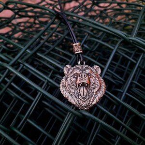 May include: A silver bear head pendant on a black leather cord with a silver bead. The pendant is hanging on a green wire fence.