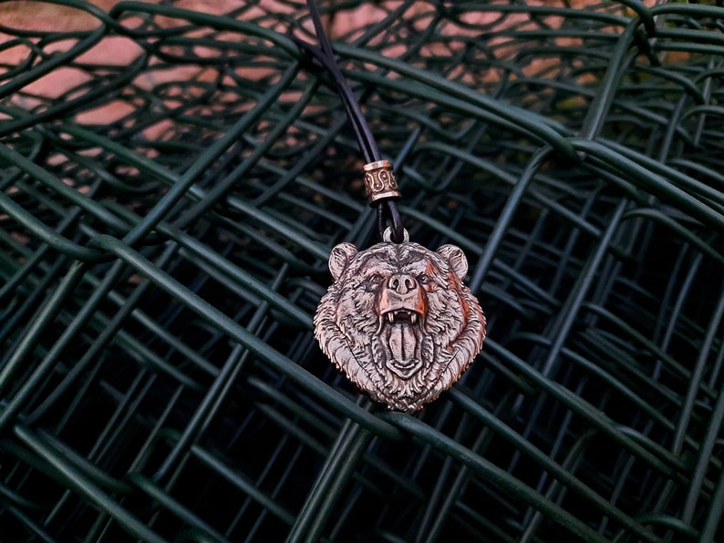 May include: A silver bear head pendant on a black leather cord with a silver bead. The pendant is hanging on a green wire fence.
