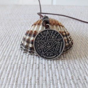 May include: A silver pendant necklace with a five-pointed star design and the words "123 TRAGRAM" around the edge. The pendant is on a brown leather cord and is resting on a seashell.