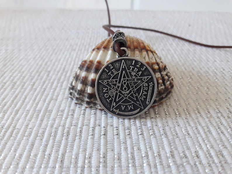 May include: A silver pendant necklace with a five-pointed star design and the words "123 TRAGRAM" around the edge. The pendant is on a brown leather cord and is resting on a seashell.