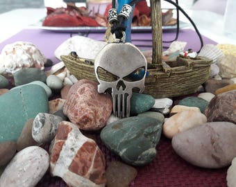 Pode incluir: Um colar com um pingente de caveira de prata com um cordão preto. O colar está sobre uma mesa com pequenas pedras e um barco de madeira em miniatura.