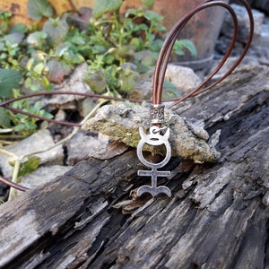 May include: A silver pendant necklace with a symbol of the planet Uranus on a brown leather cord. The necklace is lying on a piece of weathered wood.