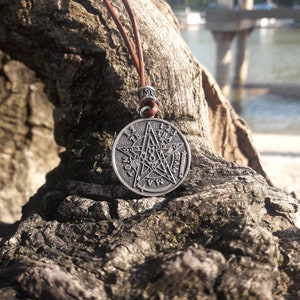 May include: A silver pendant necklace with a five-pointed star design. The star has a circle around it with numbers and symbols. The necklace is on a brown leather cord and is resting on a piece of tree bark.