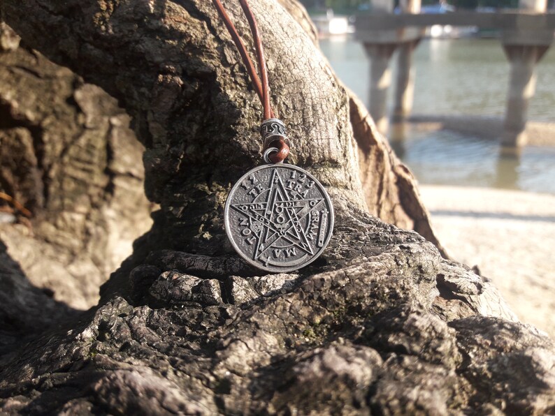 May include: A silver pendant necklace with a five-pointed star design. The star has a circle around it with numbers and symbols. The necklace is on a brown leather cord and is resting on a piece of tree bark.