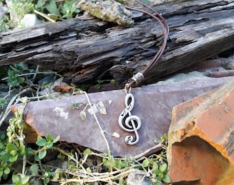 Puede incluir: Collar con colgante de clave de sol plateada y cordón de cuero marrón. El colgante detallado se destaca sobre un fondo de madera y ladrillo desgastados.