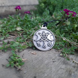 Puede incluir: Un colgante circular de metal plateado en un cordón negro. El colgante presenta un pentagrama central rodeado por dos lunas crecientes a los lados, con tres símbolos en forma de llama debajo del pentagrama y una forma de tridente arriba.