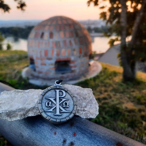 May include: A silver pendant with a Christian symbol, the Chi Rho, surrounded by a circle with the Greek letters alpha and omega. The pendant is on a rock with a blurred background of a brick dome and a river.