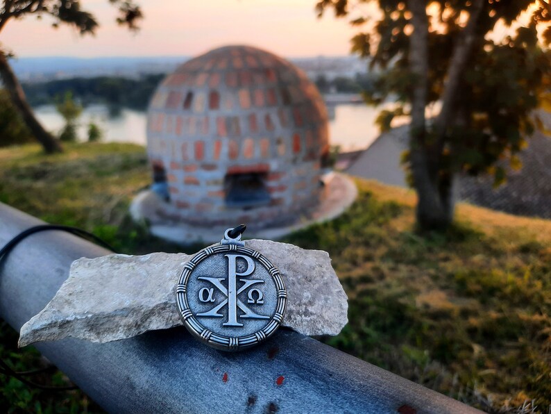 May include: A silver pendant with a Christian symbol, the Chi Rho, surrounded by a circle with the Greek letters alpha and omega. The pendant is on a rock with a blurred background of a brick dome and a river.
