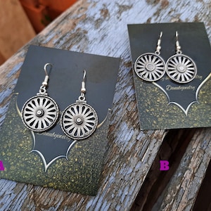 Può includere: Due paia di orecchini d'argento con un design circolare che presenta un cerchio centrale e raggi che irradiano. Gli orecchini sono esposti su carte nere con accenti dorati e il testo "Danubejewelry".