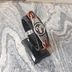 May include: Brown leather bracelet with a silver charm featuring the zodiac symbol for Aries.
