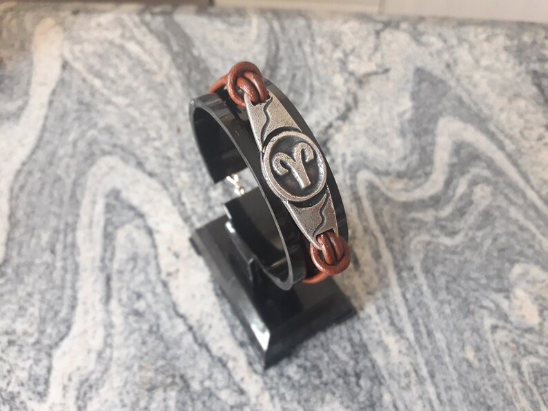 May include: Brown leather bracelet with a silver charm featuring the zodiac symbol for Aries.