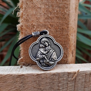 May include: A silver pendant necklace with a black leather cord. The pendant features a detailed image of a woman holding a child. The pendant is shaped like a four-leaf clover.