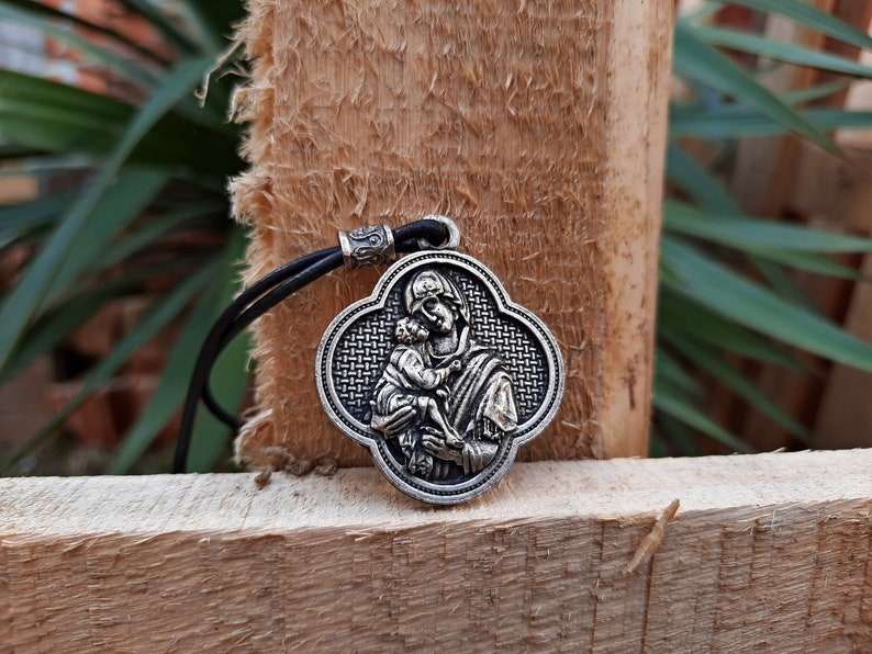 May include: A silver pendant necklace with a black leather cord. The pendant features a detailed image of a woman holding a child. The pendant is shaped like a four-leaf clover.